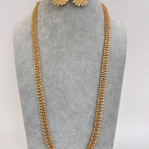 May include: A gold-coloured jewellery set comprising a long necklace and matching earrings. The necklace features a double strand design with small, round beads and teardrop-shaped accents. The earrings are circular with a floral pattern and a cluster of beads at the top.