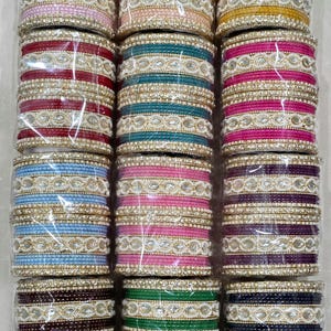 Op de afbeelding: Een verzameling kleurrijke armbanden in verschillende tinten, waaronder roze, blauw, groen en goud. Elke armband is versierd met decoratieve versieringen en gerangschikt in stapels. De armbanden zijn verpakt in doorzichtig plastic.