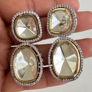 May include: Four pairs of earrings with clear, faceted center stones. Each earring has a gold-colored border and is surrounded by a halo of small, clear stones. Two pairs are square, and two are oval-shaped.