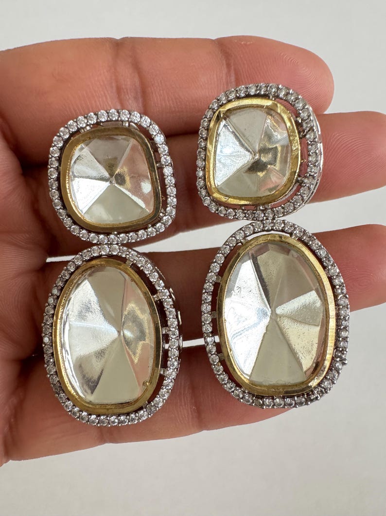 May include: Four pairs of earrings with clear, faceted center stones. Each earring has a gold-colored border and is surrounded by a halo of small, clear stones. Two pairs are square, and two are oval-shaped.