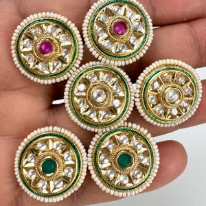 Può includere: Cinque bottoni decorativi rotondi, dorati, con perline bianche perlate attorno ai bordi. Ogni bottone ha un design centrale a forma di stella con un cerchio più piccolo al centro. I cerchi centrali presentano una gemma rosa, verde o trasparente.