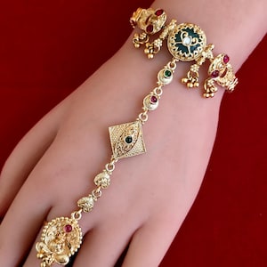May include: A gold-tone hand harness featuring a central circular medallion with a dark green inlay, accented by small red and green stones. The piece includes a delicate chain connecting the wrist band to a circular ring component with dangling beads.