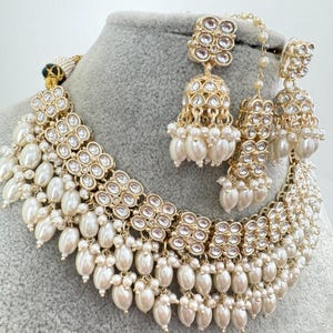 May include: A gold and white necklace and earrings set with pearl accents. The necklace features multiple layers of gold chains with clear stones and pearl drops. The earrings are dangle style with clear stones and pearl drops.