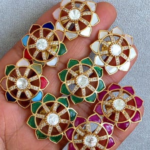 Può includere: Un gruppo di orecchini a forma di fiore color oro con una pietra centrale trasparente e pietre più piccole circostanti. I petali sono in vari colori, tra cui bianco, verde e rosso. Gli orecchini sono esposti su una superficie grigio chiaro.