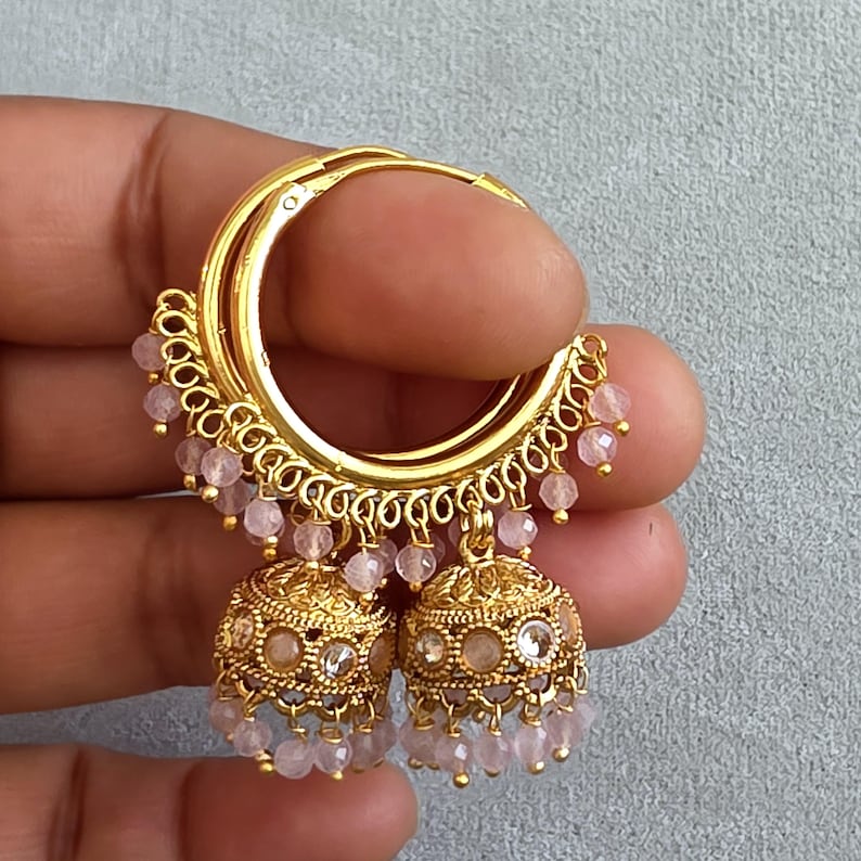 May include: Gold-toned hoop earrings with intricate filigree detailing and dangling pink beads. The earrings feature a bell-shaped charm with clear and pink accents. A stylish accessory for any occasion.