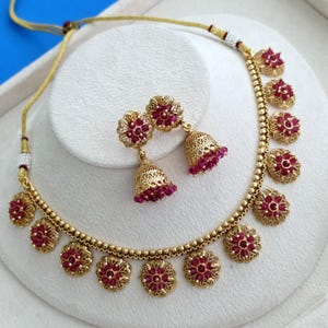 May include: A gold-toned necklace and matching jhumka earring set featuring circular floral motifs inlaid with small, deep pink stones. The necklace has a beaded chain with an adjustable cord, and the earrings have a bell-shaped drop design.