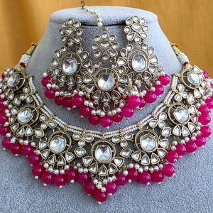 May include: A gold-toned jewelry set featuring a wide choker necklace and matching earrings with a central maang tikka. The pieces are adorned with clear, teardrop-shaped stones, small white seed pearls, and vibrant fuchsia pink spherical beads.