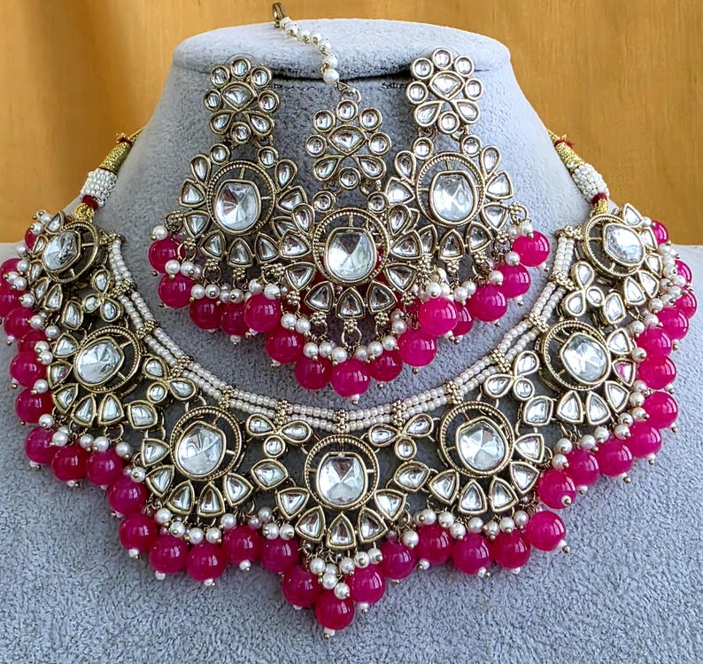 May include: A gold-toned jewelry set featuring a wide choker necklace and matching earrings with a central maang tikka. The pieces are adorned with clear, teardrop-shaped stones, small white seed pearls, and vibrant fuchsia pink spherical beads.