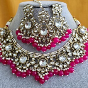 May include: A gold-toned jewelry set consisting of a necklace, a pair of earrings, and a maang tikka. Each piece features clear, teardrop-shaped stones set in metal, accented with small white beads and dangling bright pink spherical beads.