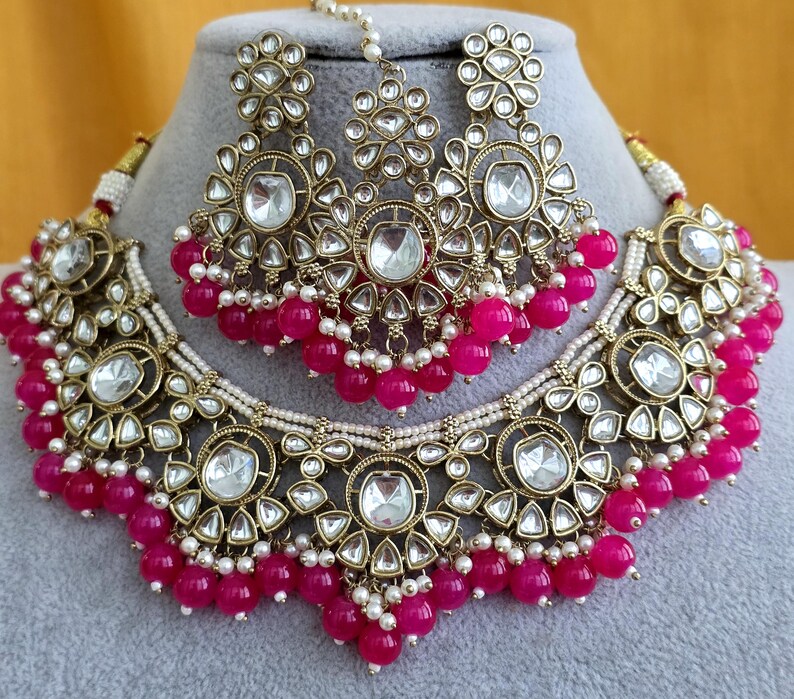 May include: A gold-toned jewelry set consisting of a necklace, a pair of earrings, and a maang tikka. Each piece features clear, teardrop-shaped stones set in metal, accented with small white beads and dangling bright pink spherical beads.