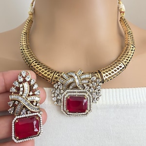 May include: A gold-coloured necklace and earring set featuring large, rectangular, ruby-red gemstones. The necklace has a geometric design with a central pendant and matching earrings. The earrings are held by a hand. The jewellery is set with clear, sparkling stones.
