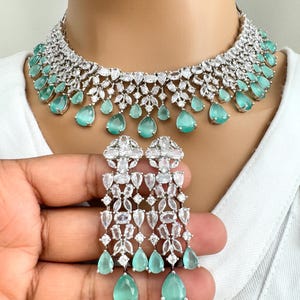 Op de afbeelding: Een zilveren en witte ketting en oorbellenset met druppelvormige groene stenen. De ketting heeft een gelaagd ontwerp met een rij grotere stenen aan de onderkant. De oorbellen zijn lang en hangen.