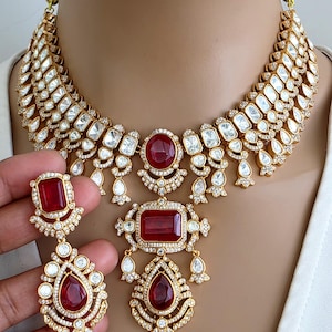 May include: A gold-toned necklace and matching earring set featuring deep red rectangular and pear-shaped stones. The jewelry is embellished with numerous small white stones set in intricate metalwork, creating a layered, ornate design.
