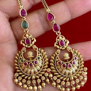 May include: A pair of gold-toned dangle earrings featuring a chain-link top, teardrop-shaped pink and green stones, and a crescent-shaped pendant base. The base is decorated with small floral motifs, embedded pink and green stones, and dangling beads.