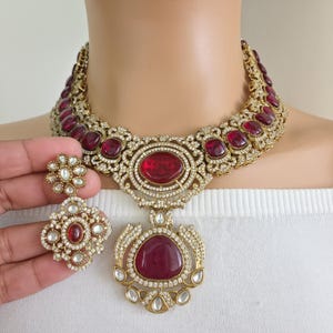May include: A gold-toned necklace and earring set featuring large, oval and teardrop-shaped red gemstones. The necklace has a bib design with smaller red stones and clear crystals. The earrings have a floral design with a central red stone.