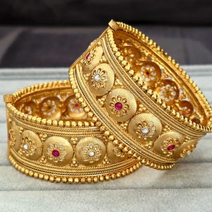 May include: Two gold-toned bangles featuring a repeating pattern of circular medallions with textured surfaces. Each medallion is centered with a small floral motif containing either a red or clear stone. The edges are lined with beaded detailing.