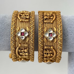 May include: A pair of gold-toned bangles with intricate designs and red and white gemstones. The bangles are decorated with small, raised circles and floral motifs.