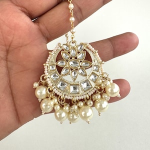 May include: Gold and white beaded maang tikka with a round, ornate design featuring clear stones. The maang tikka has a chain and a pearl drop.