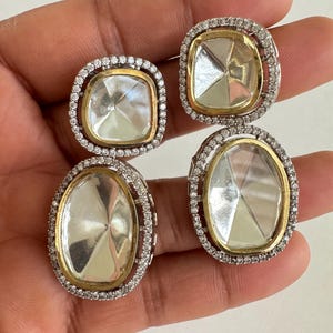 May include: A pair of silver and gold-toned earrings. Each earring features a large, clear, faceted stone set within a gold frame, surrounded by a halo of small, clear stones. The earrings have a square and oval shape.