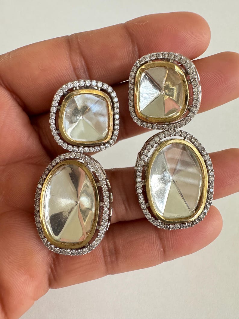 May include: A pair of silver and gold-toned earrings. Each earring features a large, clear, faceted stone set within a gold frame, surrounded by a halo of small, clear stones. The earrings have a square and oval shape.