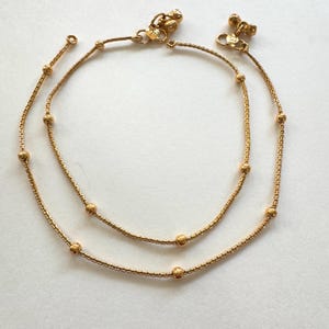 K&ouml;nnte beinhalten: Ein Paar goldene Fu&szlig;kettchen mit kleinen runden Perlen. Die Fu&szlig;kettchen bestehen aus einer zarten Kette und haben ein schlichtes Design.