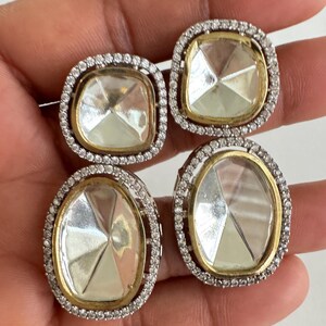 May include: Four square and oval-shaped earrings with clear, faceted center stones. Each earring is surrounded by a gold-toned metal frame and a halo of small, clear crystals. The earrings are set against a neutral background.