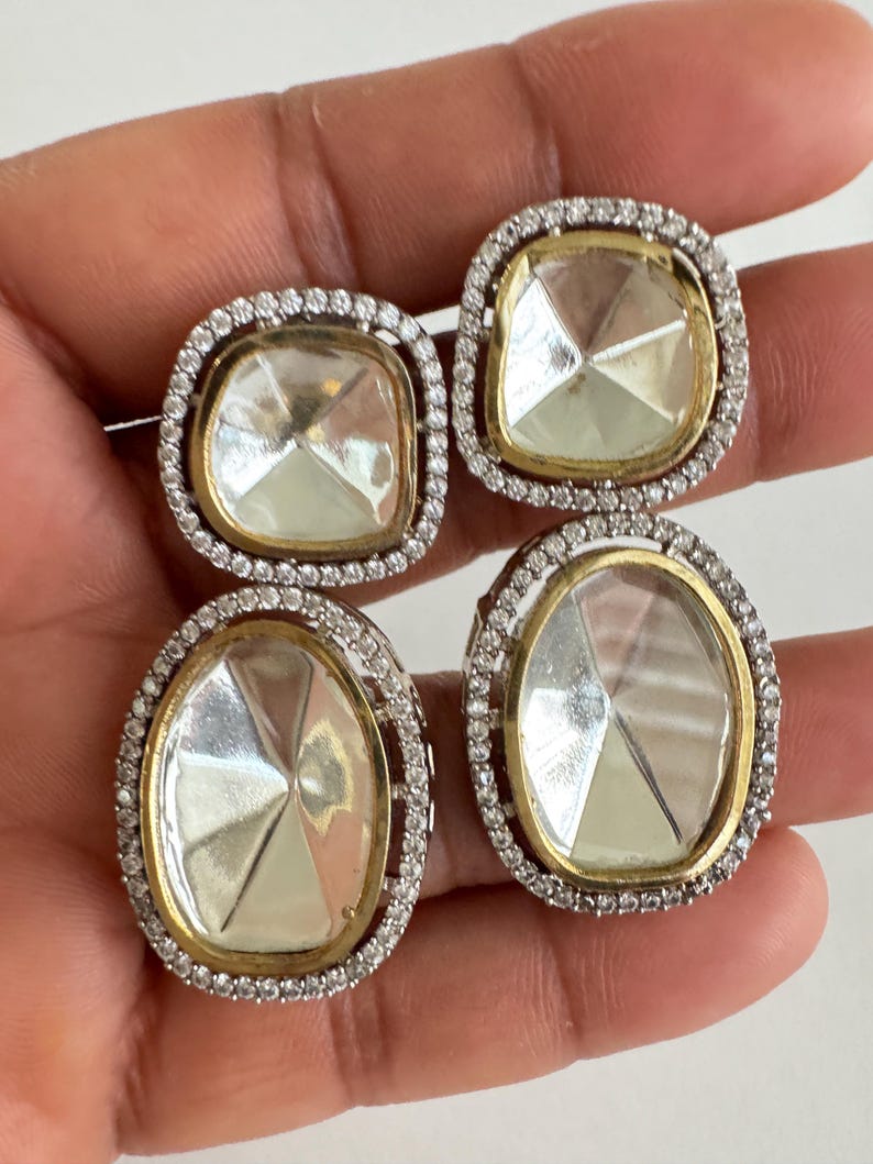 May include: Four square and oval-shaped earrings with clear, faceted center stones. Each earring is surrounded by a gold-toned metal frame and a halo of small, clear crystals. The earrings are set against a neutral background.