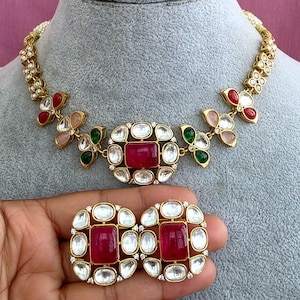 Peut inclure: Un collier et des boucles d'oreilles assorties en métal doré, ornés d'une pierre rouge rectangulaire centrale entourée de pierres blanches. Le collier présente des motifs floraux avec des pierres rouges, vertes et rose clair serties en métal doré.