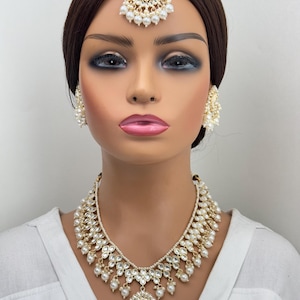 May include: A gold and white Indian bridal jewelry set with a necklace, earrings, and a tikka. The necklace features a teardrop pendant with pearls and stones. The earrings are round with pearls and stones. The tikka is a traditional forehead ornament with pearls and stones.