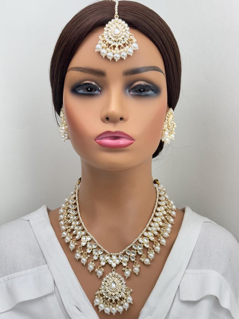 May include: A gold and white Indian bridal jewelry set with a necklace, earrings, and a tikka. The necklace features a teardrop pendant with pearls and stones. The earrings are round with pearls and stones. The tikka is a traditional forehead ornament with pearls and stones.