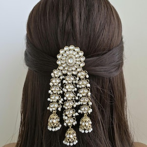 Puede incluir: Accesorio para el cabello dorado con un diseño floral y elementos colgantes. El accesorio presenta piedras claras y brillantes y pequeñas perlas blancas. Tres colgantes en forma de campana cuelgan de la parte inferior, agregando movimiento y detalle.