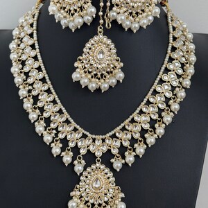 May include: A gold necklace and earrings set with white pearl accents and a teardrop-shaped pendant. The necklace features a layered design with multiple strands of pearls and intricate floral motifs.