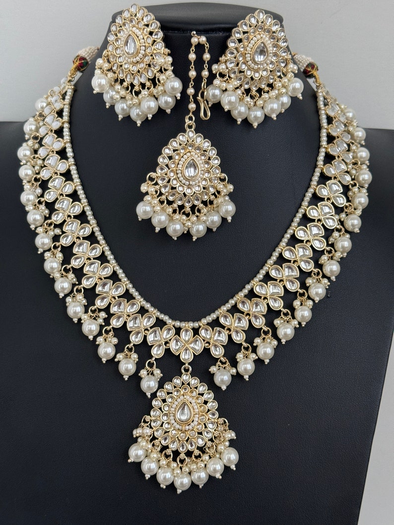 May include: A gold necklace and earrings set with white pearl accents and a teardrop-shaped pendant. The necklace features a layered design with multiple strands of pearls and intricate floral motifs.