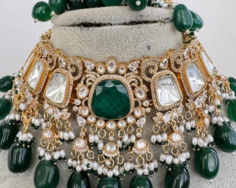 May include: An ornate gold necklace with emerald green and clear gemstones. The necklace features a large, square emerald center stone, surrounded by smaller clear stones and dangling emerald drops. White pearls accent the design.