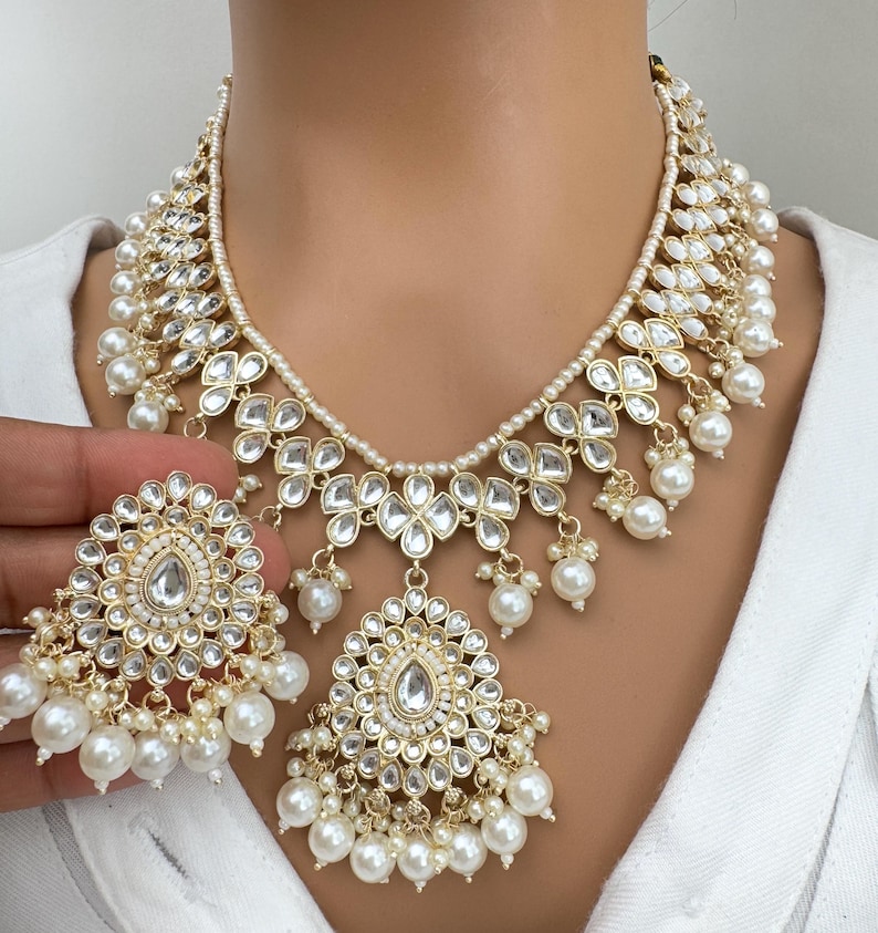 May include: A gold necklace with white pearl accents and a matching pair of earrings. The necklace features a large, ornate pendant with a teardrop-shaped center stone.