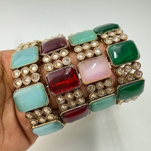 Puede incluir: Cuatro pulseras de oro con piedras rectangulares de colores y brillantes transparentes. Las pulseras están apiladas una encima de la otra. Las piedras son turquesa, roja, rosa y verde.