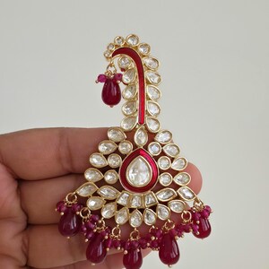 May include: A decorative gold earring with red enamel and clear stones. The earring has a teardrop shape with dangling red beads. A unique piece of jewellery.