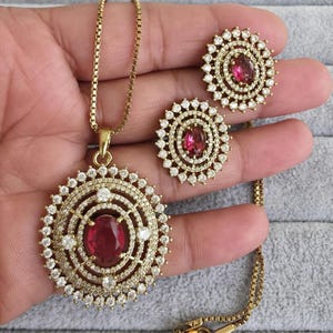 Może przedstawiać: Złoty zestaw biżuterii składający się z naszyjnika z wisiorkiem i pasujących kolczyków. Wisiorek i kolczyki mają centralny owalny rubinowo-czerwony kamień otoczony przez halo małych, przezroczystych kryształków. Naszyjnik ma delikatny łańcuszek.