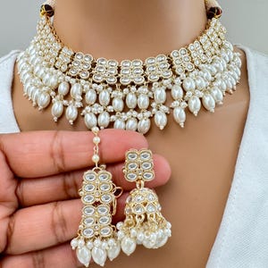 May include: A gold and white choker necklace with pearl accents and matching earrings. The necklace has a layered design with multiple rows of pearls and sparkling stones.