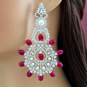 May include: A large chandelier earring featuring a silver-toned metal frame encrusted with clear, marquise-cut crystals. The design includes a central white pearl, smaller accent pearls, and several teardrop-shaped, deep red gemstones.