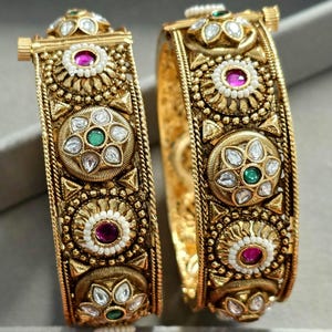 Pode incluir: Dois braceletes dourados com dobradiça, apresentando um trabalho de metal intrincado, motivos florais circulares e pedras incrustadas. O design inclui pedras centrais redondas rosas e verdes alternadas, rodeadas por contas brancas e pétalas claras.