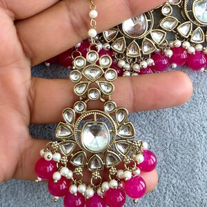 May include: A gold-toned maang tikka featuring clear, teardrop-shaped stones arranged in a floral pattern, accented with small white pearls and dangling bright pink spherical beads. The piece includes a delicate chain with white pearl accents.