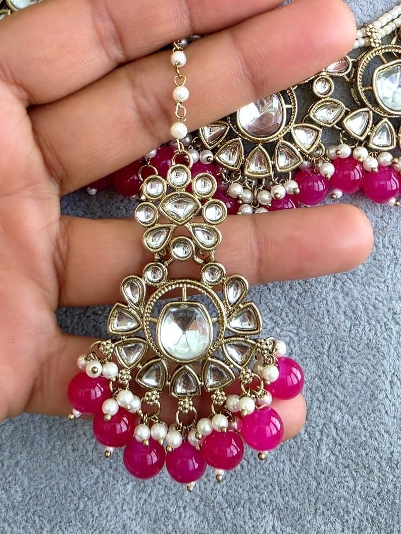 May include: A gold-toned maang tikka featuring clear, teardrop-shaped stones arranged in a floral pattern, accented with small white pearls and dangling bright pink spherical beads. The piece includes a delicate chain with white pearl accents.