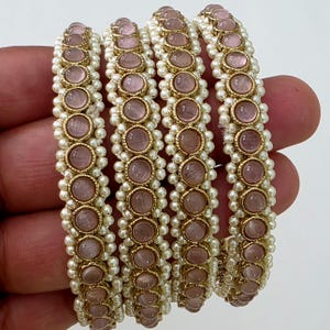 May include: Four gold-toned bangles with alternating pink gemstones and small white pearls. The gemstones are set in gold bezels, and the pearls create a border around the gemstones. The bangles are held in a hand.
