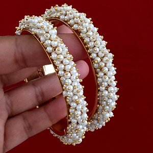 May include: Two circular gold-toned bangles featuring a dense, textured surface covered in small, round white faux pearls. The interior of each bangle is smooth and metallic gold, providing a contrast to the beaded exterior surface.