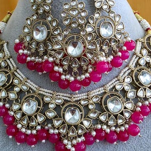 May include: A gold-toned jewelry set featuring a necklace and a maang tikka, both adorned with clear, teardrop-shaped stones set in intricate metalwork. The pieces are accented with small white seed pearls and vibrant fuchsia pink beads.