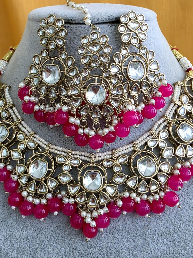 May include: A gold-toned jewelry set featuring a necklace and a maang tikka, both adorned with clear, teardrop-shaped stones set in intricate metalwork. The pieces are accented with small white seed pearls and vibrant fuchsia pink beads.