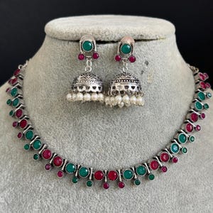 May include: A silver necklace with green and pink stones, and matching earrings with pearl accents. The necklace has a delicate design with alternating green and pink stones.