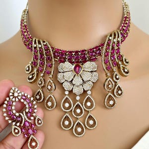 May include: A luxurious necklace and earring set featuring a floral pendant and teardrop dangles. The jewelry is adorned with ruby-colored stones and clear crystals, set in a gold-toned metal. The necklace is a statement piece.