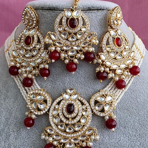 Può includere: Un set di gioielli dorati ornato con una collana, orecchini e un copricapo. I pezzi sono adornati con pietre preziose rosse, cristalli trasparenti e piccole perle bianche. La collana ha più fili di perline bianche.